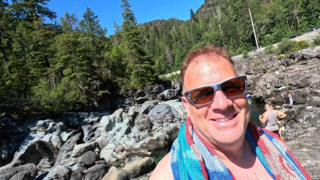 Me at ?a?ukmin Tribal Park, a spot also known as Wally Creek. I've got sunglasses on with a towel around my neck and some people and waterfalls behind me.